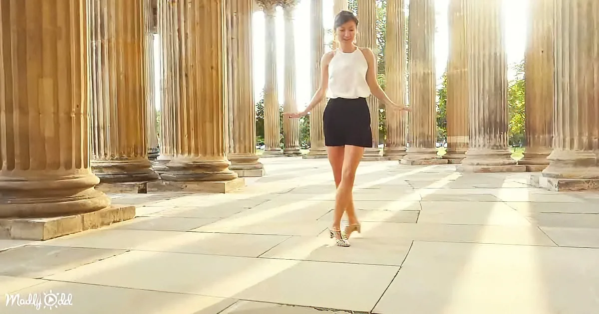 A woman in a white top and black skirt walks gracefully among tall, sunlit columns.