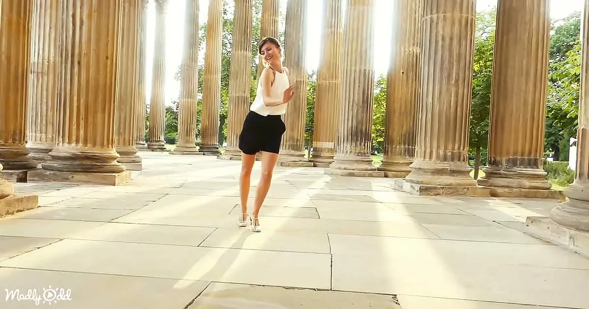 A woman in a white top and black skirt stands gracefully in front of tall, classical columns on a sunny day.
