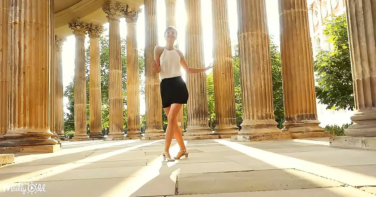 A woman in a white top and black skirt dances joyfully among tall, sunlit columns.