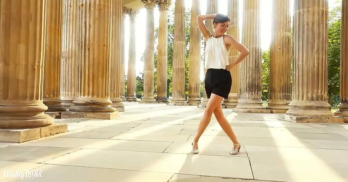 A woman in a white top and black shorts poses confidently in front of tall, sunlit columns.