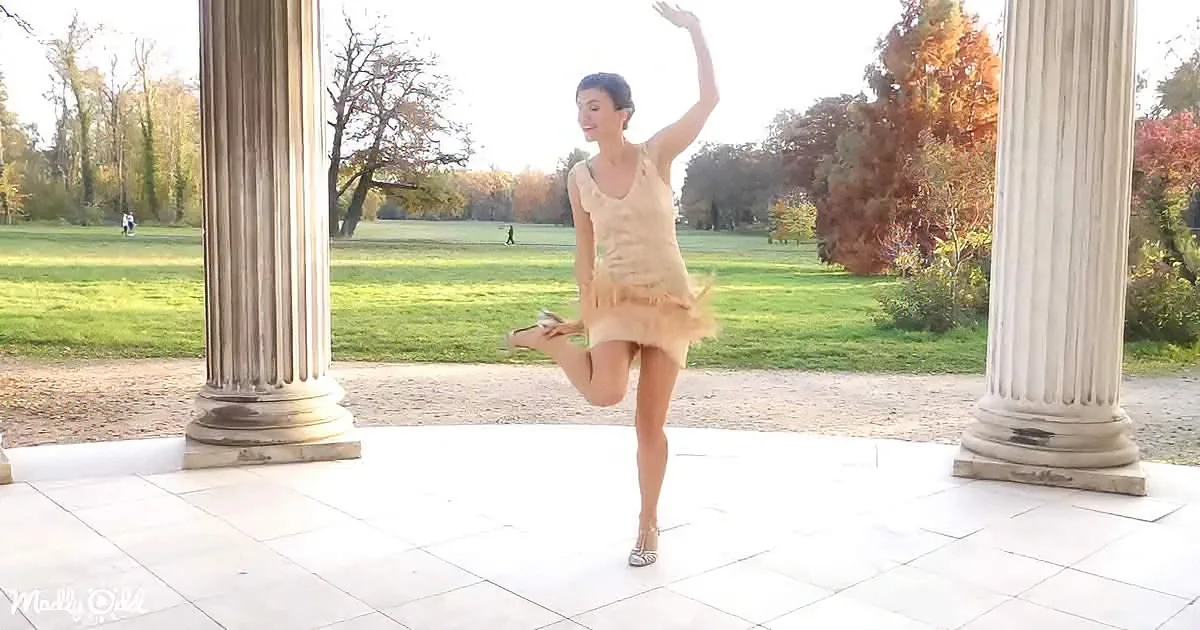 A dancer in a beige dress performs a ballet pose on a stone platform with columns, surrounded by autumn foliage.