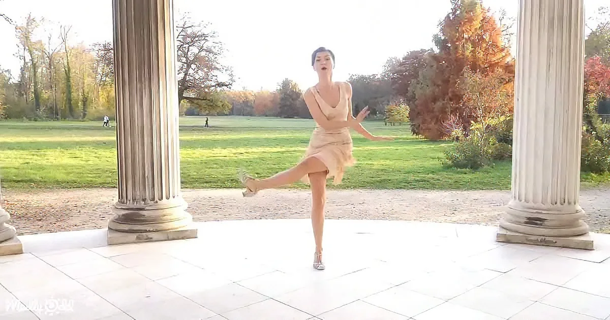A woman in a beige dress balances on one leg in a classical setting with columns and autumn foliage.