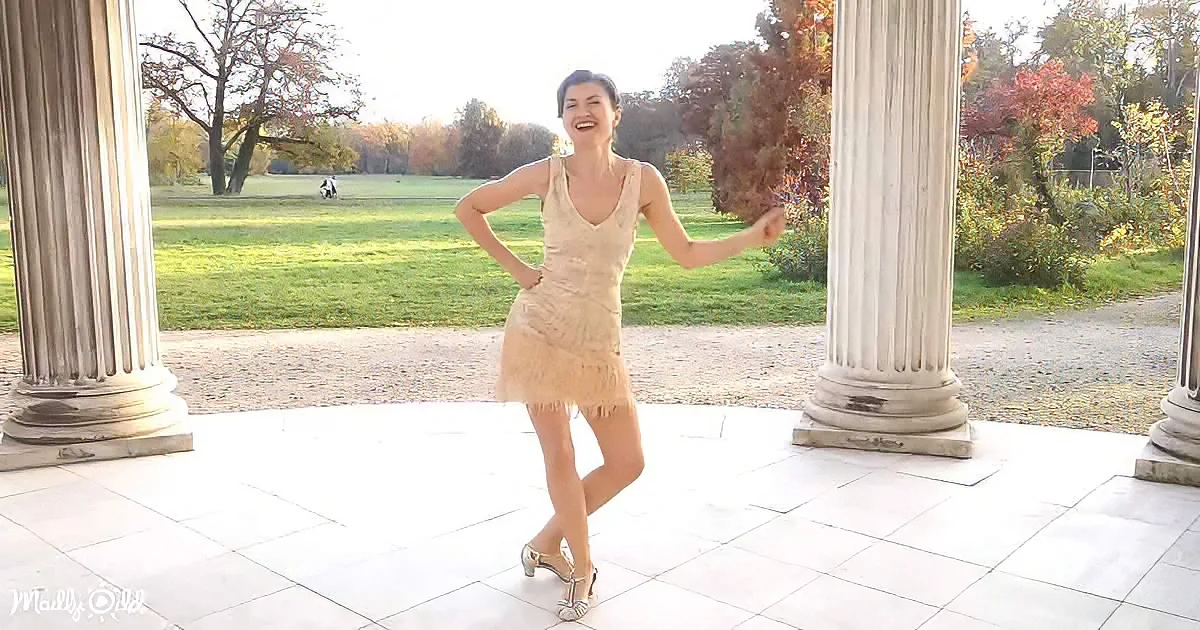 A woman in a beige dress dances joyfully between white columns in a park.