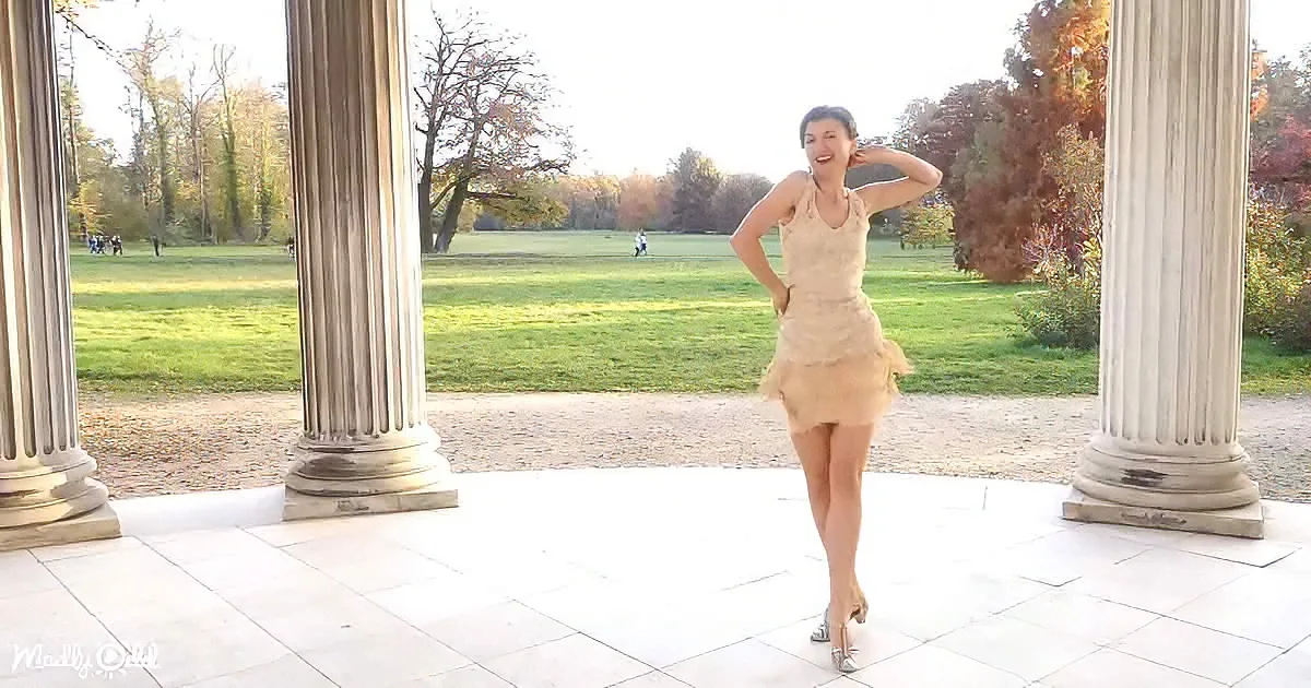 A woman in a beige dress stands on a stone platform with columns, smiling, in a park with green grass and autumn trees.