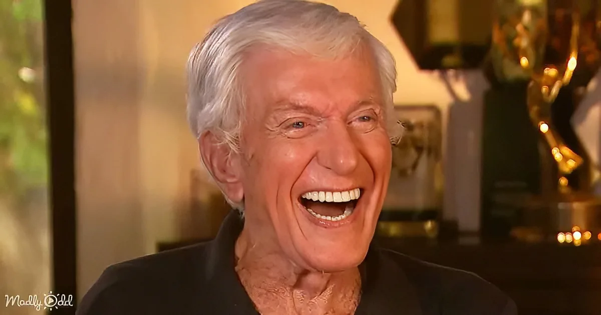 An older man with white hair smiles widely, wearing a black shirt, with blurred background and golden trophy visible.