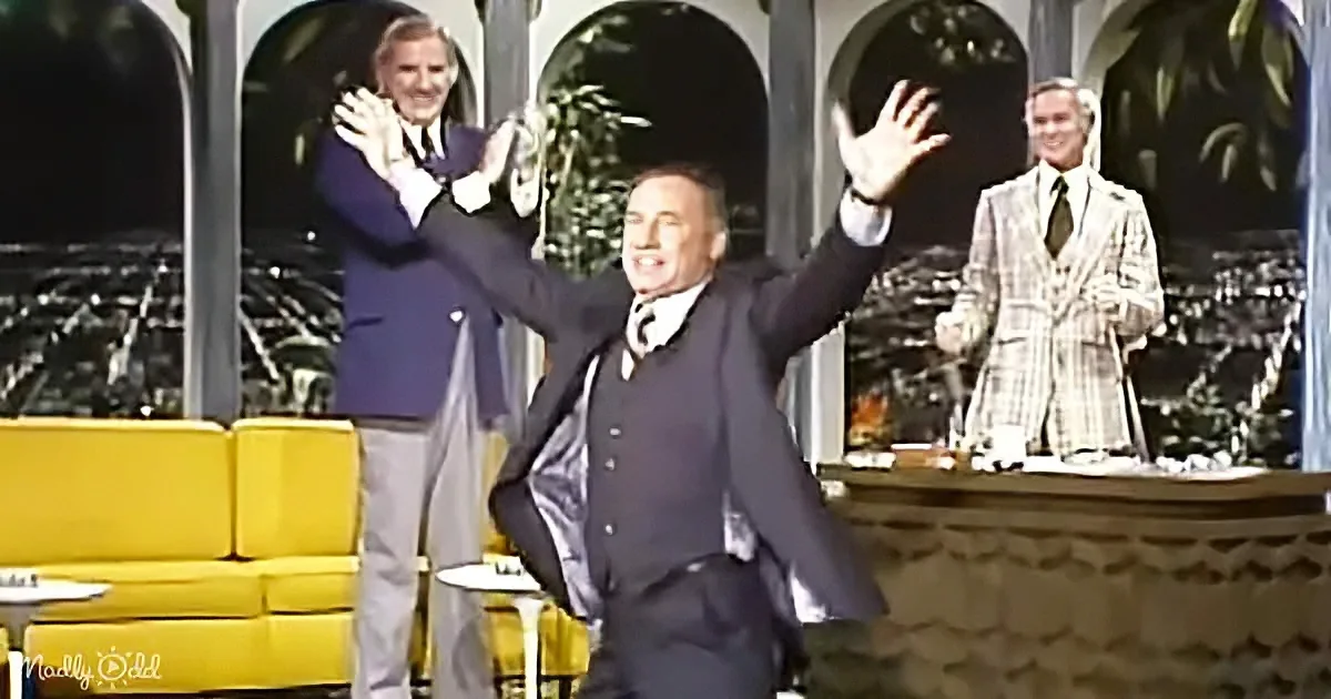 A man in a suit raises his arms in celebration on a stage with two others in the background.