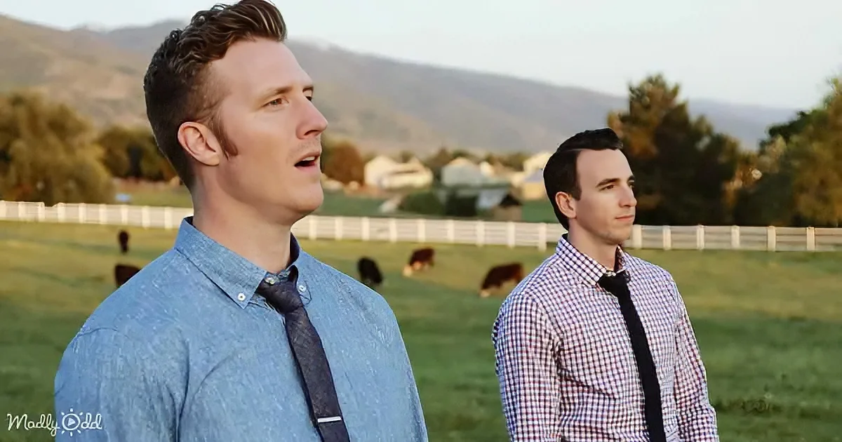 Two men in shirts and ties stand in a grassy field with cows and mountains in the background.