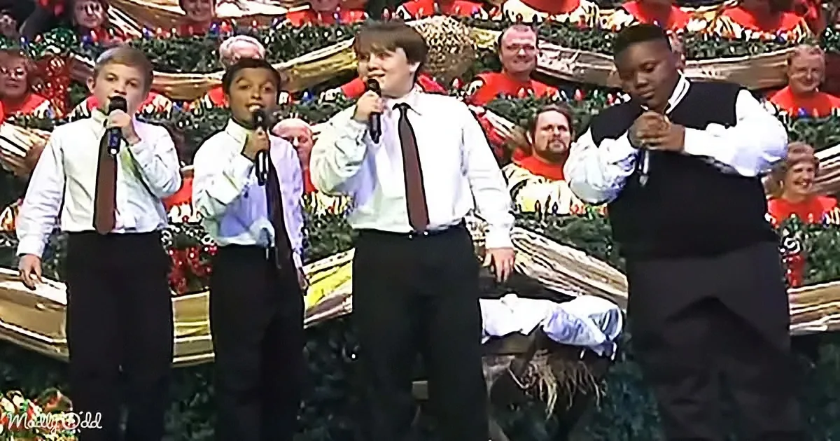 Four young men in formal attire sing into microphones on a stage with a festive background.