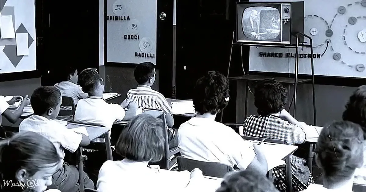 Children sit at desks watching a teacher on a small TV in a classroom.