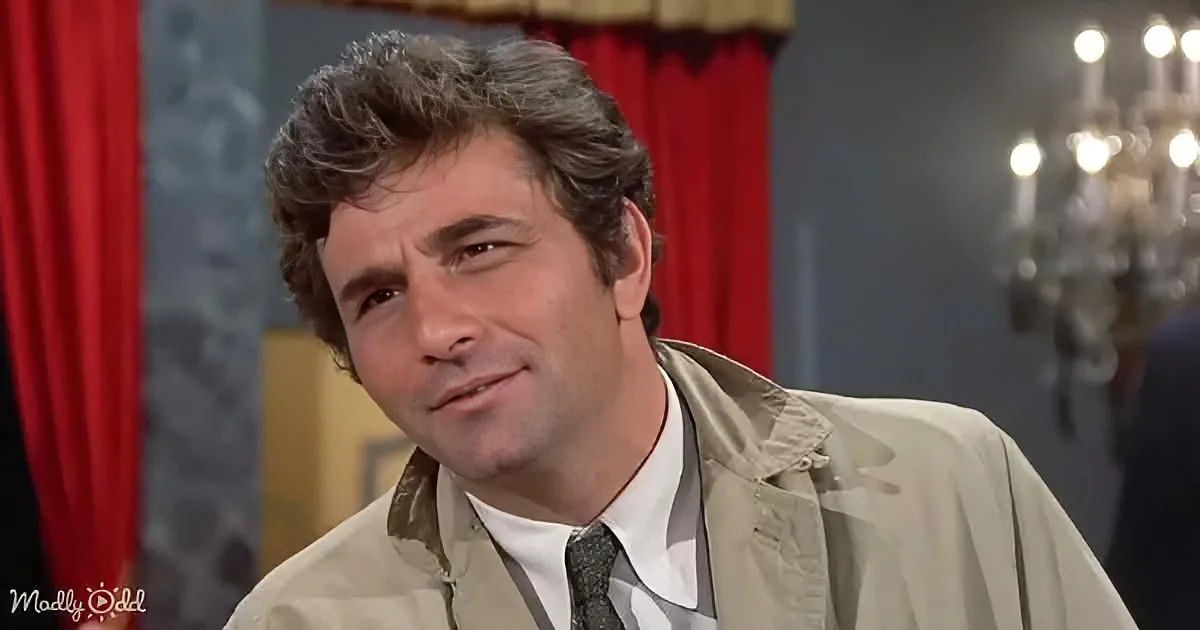 A man with curly hair, wearing a beige jacket over a white shirt and tie, smiles slightly in a room with red curtains and...