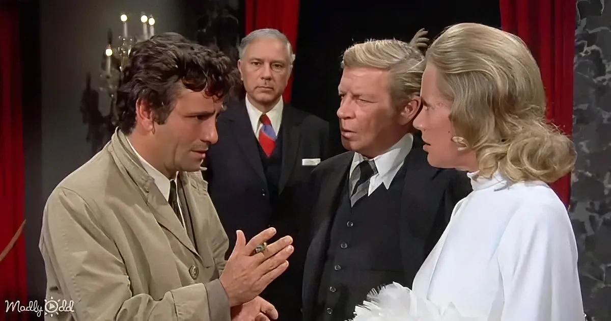 A man in a trench coat gestures while talking to a woman in white, with two men in suits observing in the background.