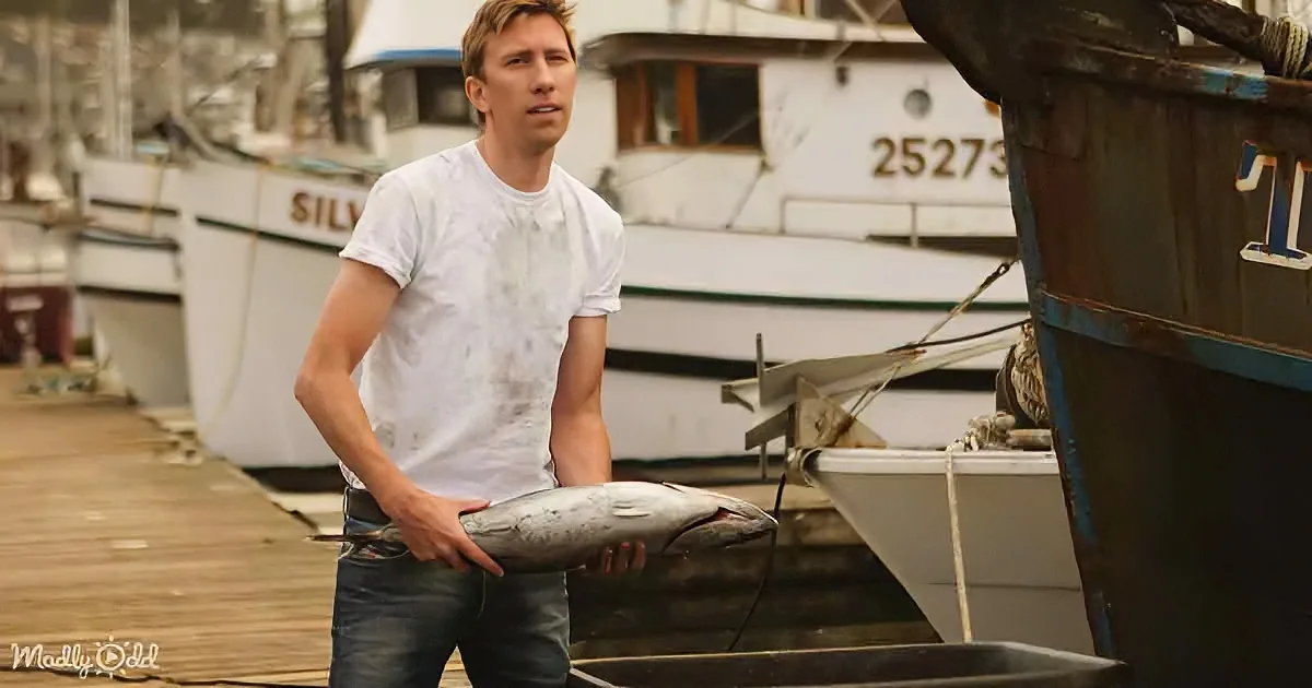 A man holds a fish on a dock with boats in the background.