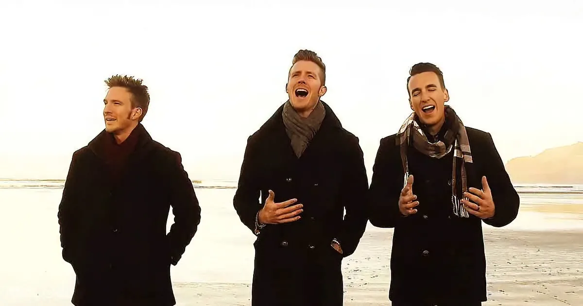 Three men in black coats and scarves sing joyfully on a snowy beach.