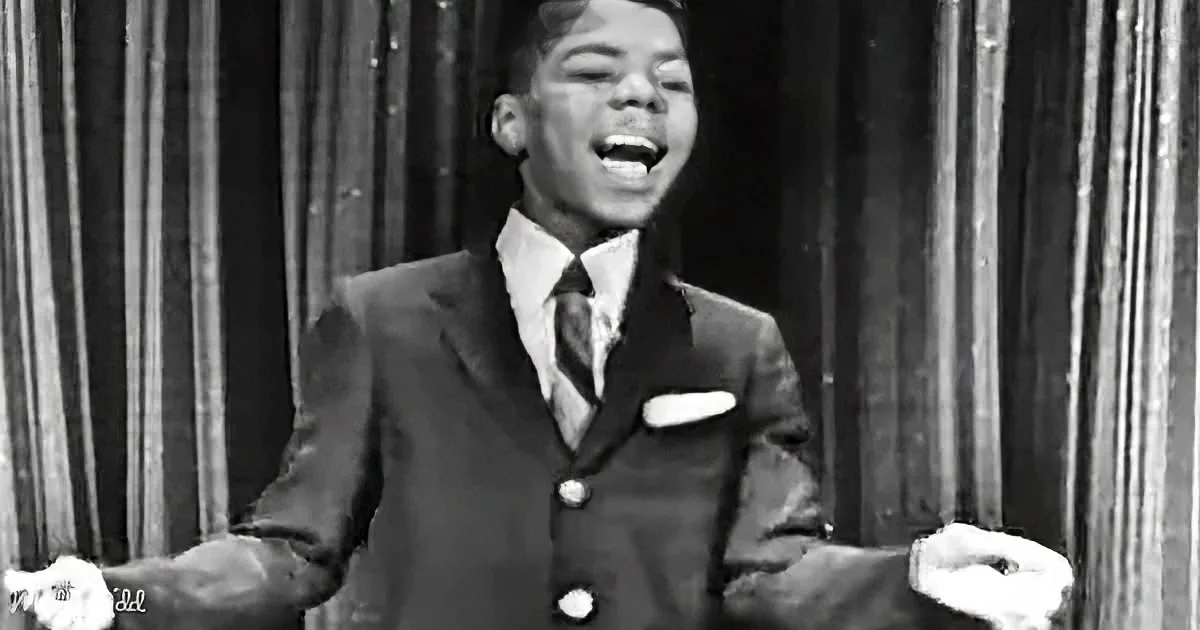 A man in a suit and tie is singing with his arms outstretched, smiling.