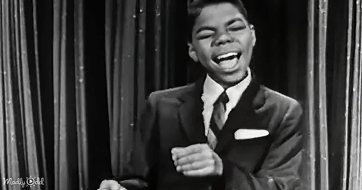 A young man in a suit and tie is singing passionately on stage, with a curtain backdrop.
