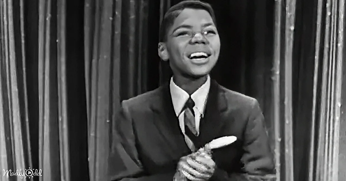 A young man in a suit and tie smiles widely, holding a microphone, against a curtain backdrop.