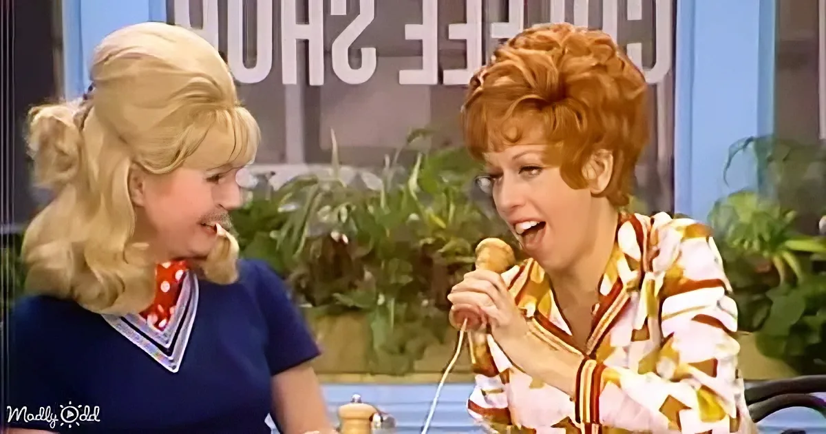 A woman with red hair holds a microphone while another with blonde hair looks on, both seated at a table.