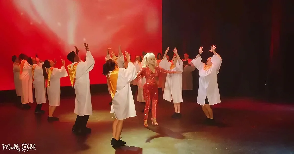 A performer in a red sequined outfit dances center stage with a group in white robes, against a red-lit backdrop.