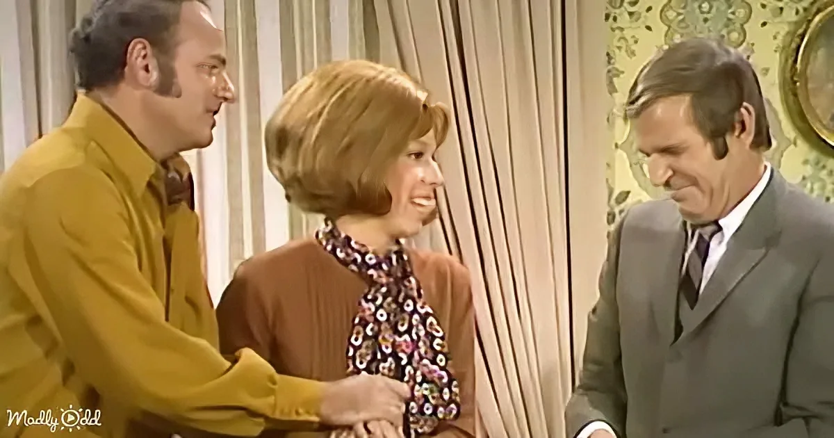 A woman in a floral blouse smiles as a man in a yellow shirt and another in a gray suit look at her.