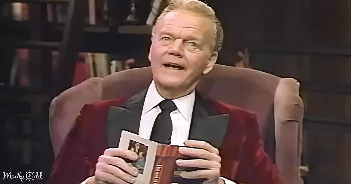 A man in a suit sits in a chair, holding a framed picture and appearing surprised or excited.