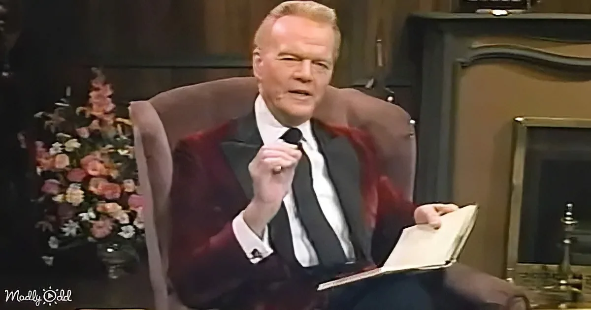 A man in a suit sits in a chair, holding a microphone and a book, with a floral arrangement and fireplace in the background.