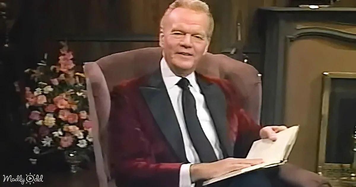 A man in a suit sits in a chair, holding an open book, with a floral arrangement and a fireplace in the background.