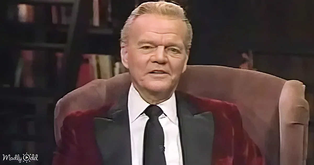 A man in a suit and tie sits in a red chair, looking serious, against a dark background.
