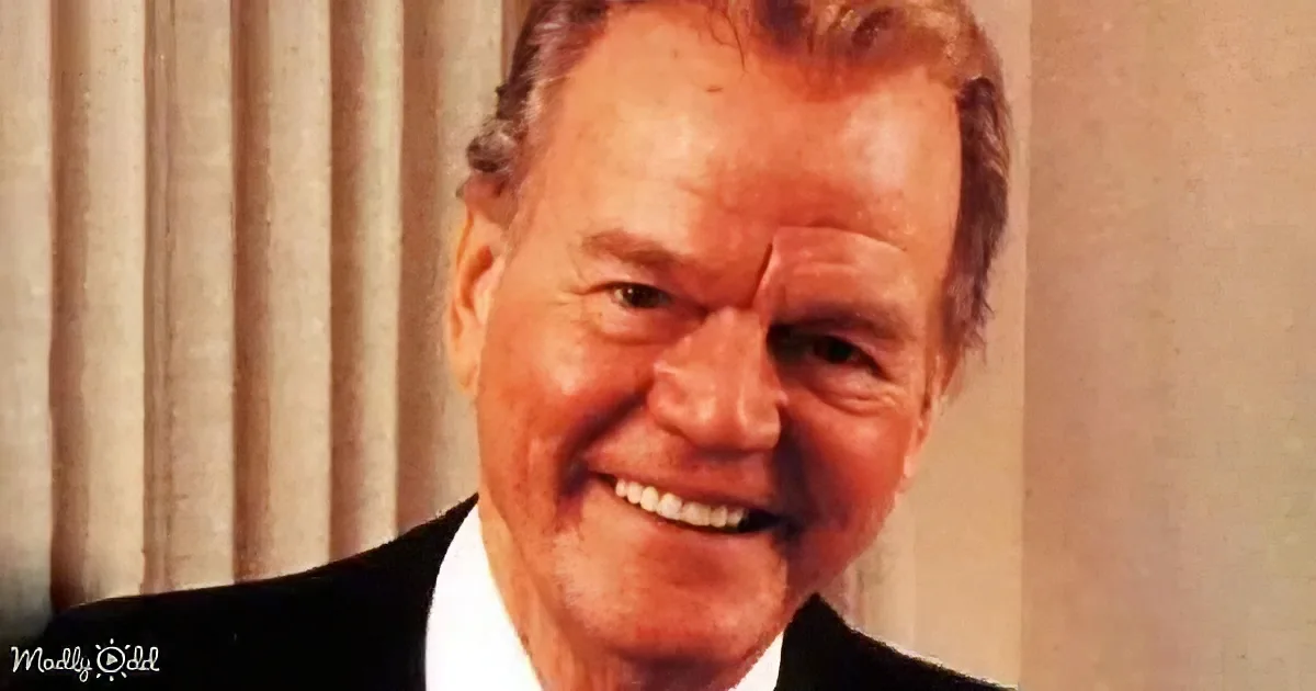A smiling man with short, styled hair, wearing a black suit and white shirt, against a beige curtain backdrop.