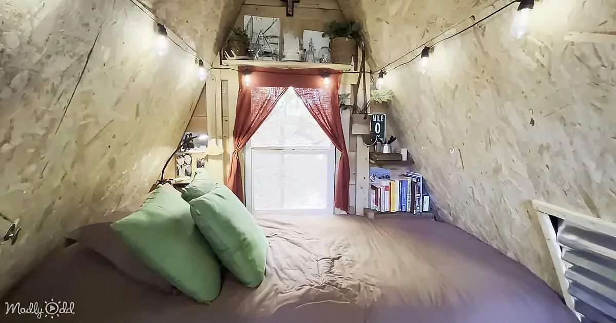 A cozy bedroom with a bed, string lights, a window with red curtains, and a bookshelf.