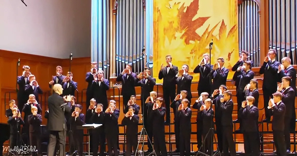 A choir in formal attire performs on stage with a conductor, surrounded by an ornate organ and autumn-themed backdrop.