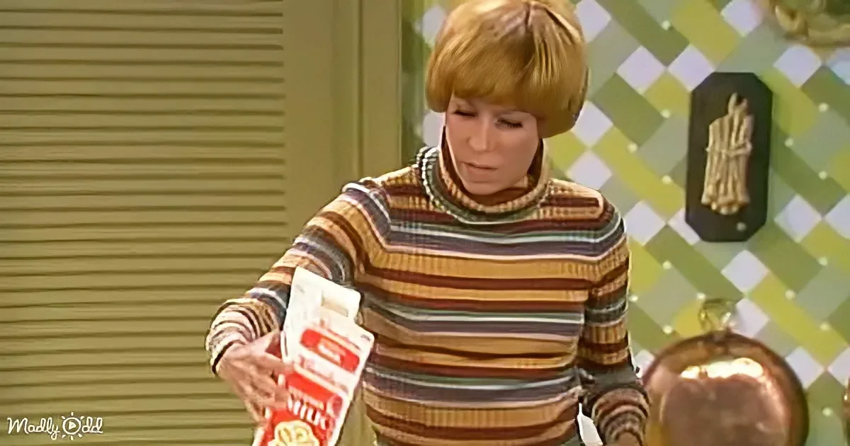 A woman in a striped sweater holds a red box, looking down, with a green checkered wall and decorative items in the...