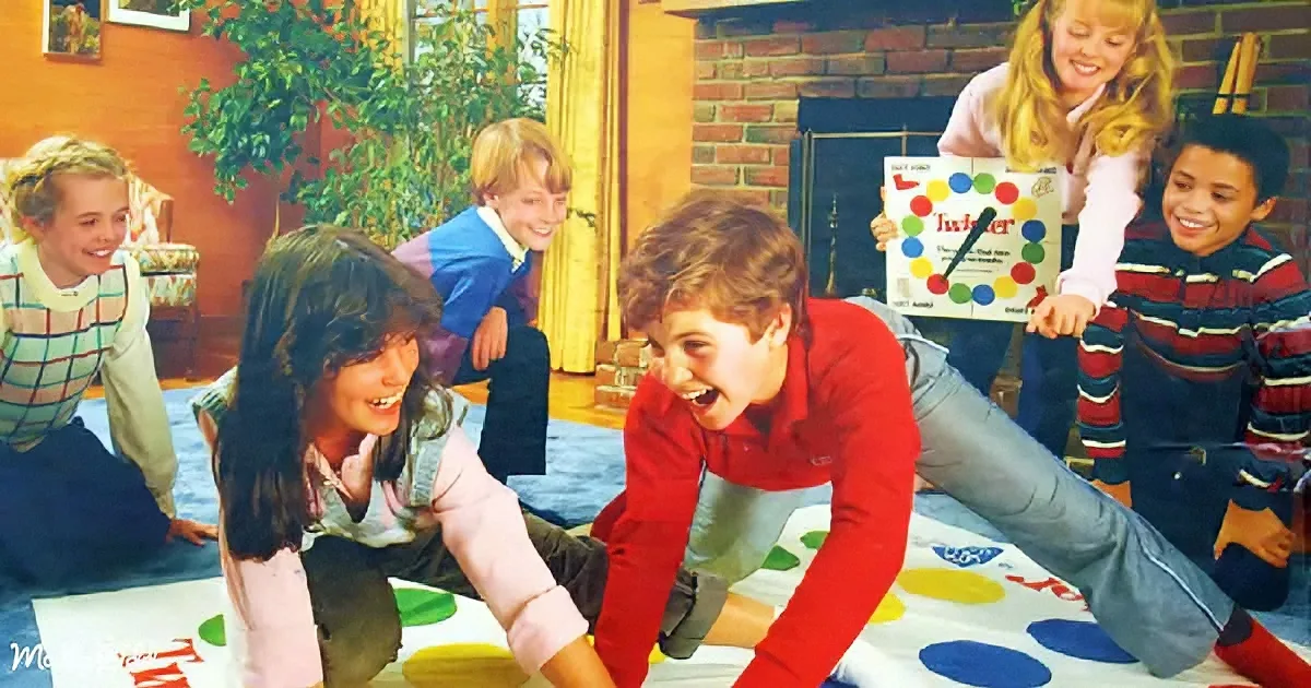 A boy in a red shirt is playfully wrestling with a girl on a Twister mat, surrounded by smiling children.
