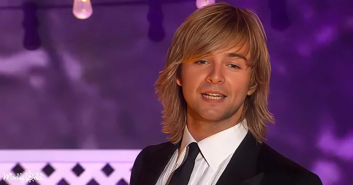 A man with long blonde hair in a suit and tie smiles against a purple-lit background.