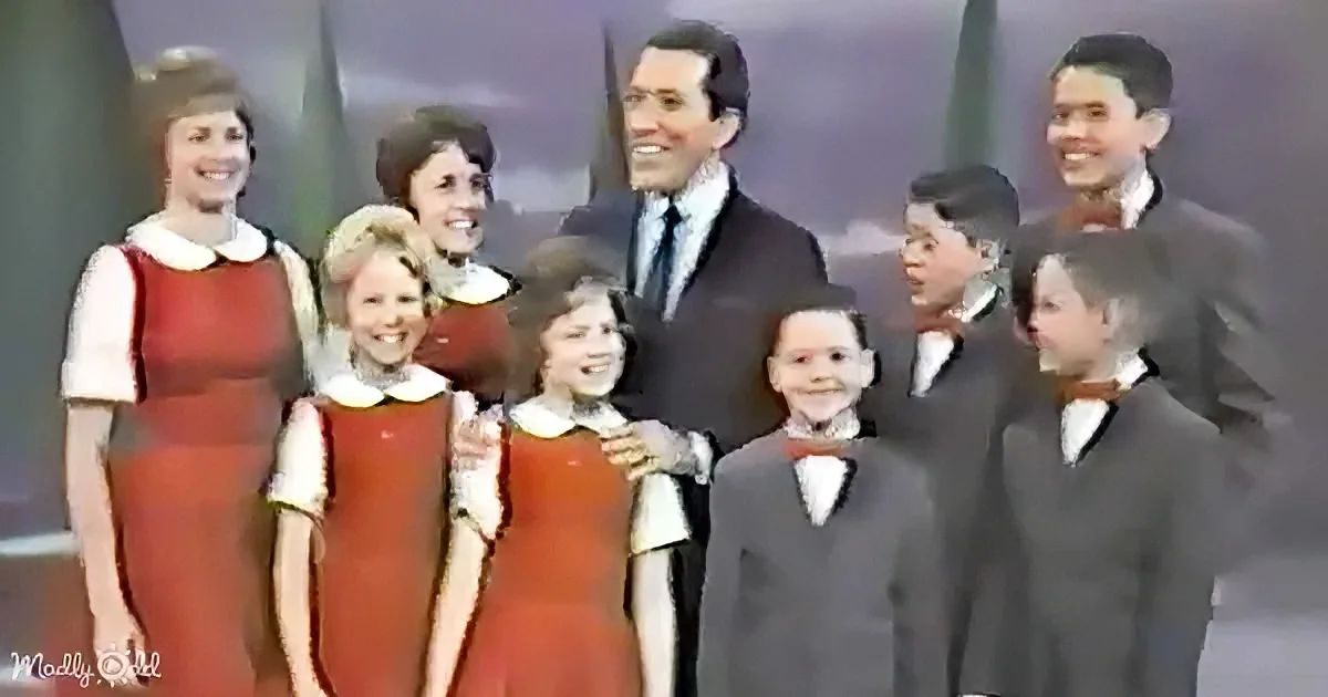 A man in a suit stands with a group of children in red and gray outfits, smiling.