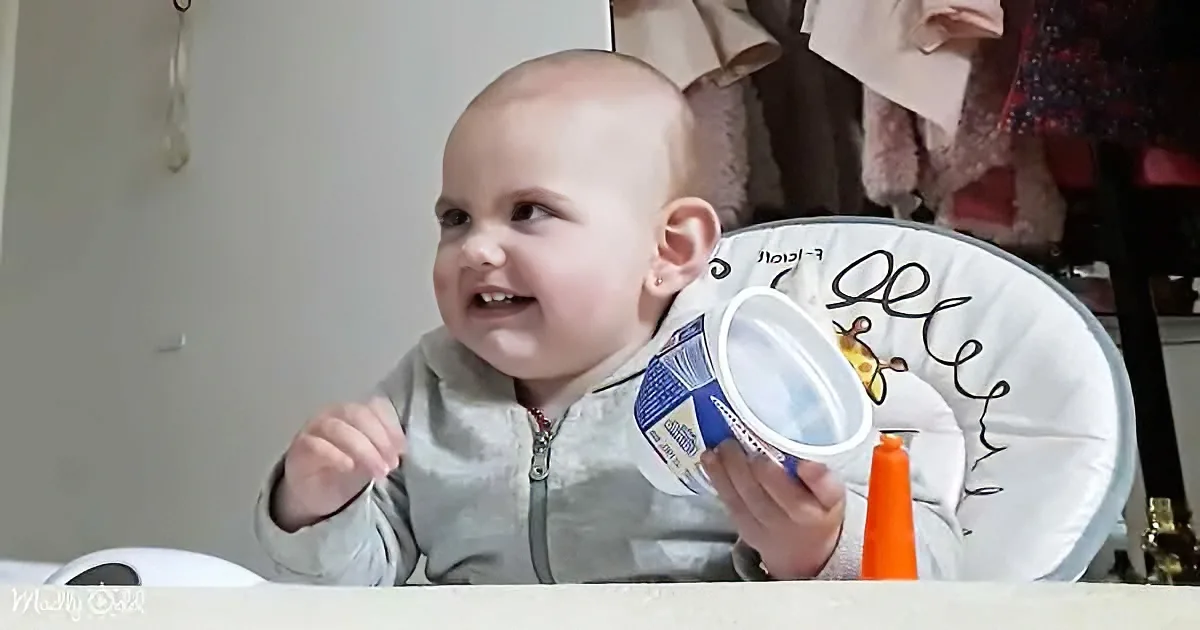 A baby in a high chair smiles while holding a container, with a colorful bib and orange cone nearby.