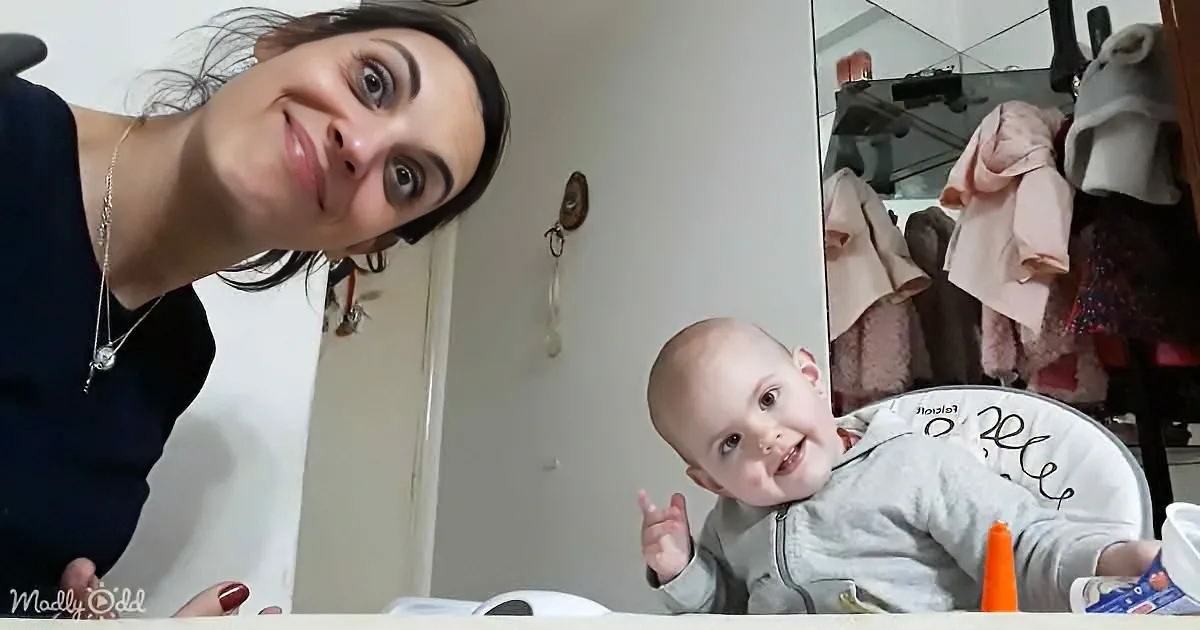 A woman leans over, smiling, with a baby in a high chair, both in a room with a mirror and clothes.