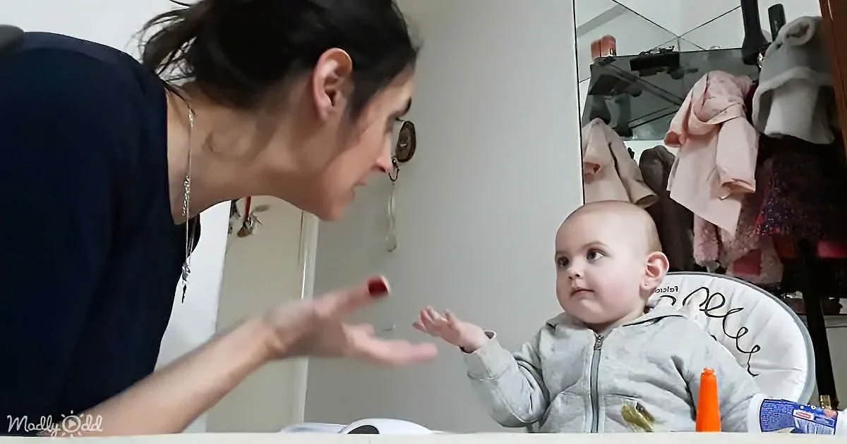A woman leans towards a baby in a high chair, gesturing with her hand.
