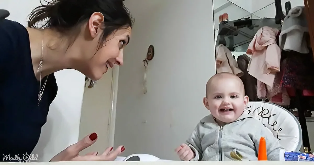 A woman leans towards a smiling baby in a high chair, both appearing happy and engaged.