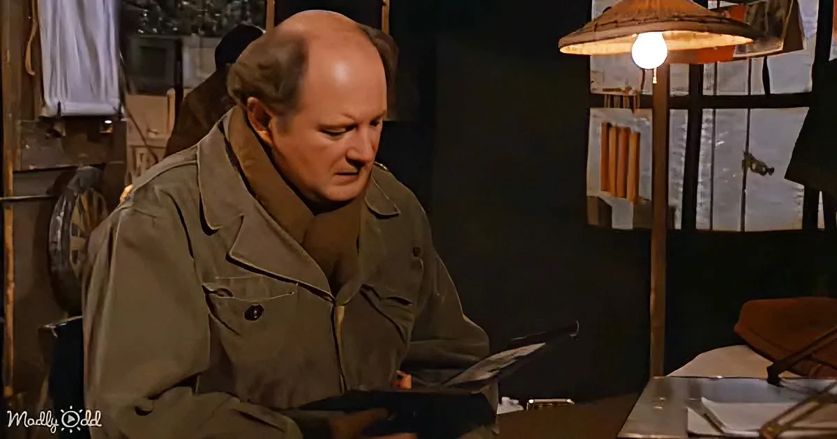 A man in a brown jacket sits at a table, reading a book under a lamp, surrounded by a cluttered room.