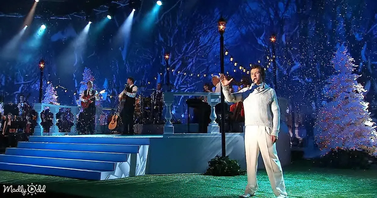 A man in white gestures on stage with a band and Christmas trees in the background.