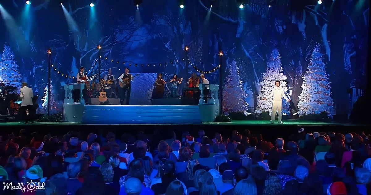 A performer in white stands on stage with a band, surrounded by a snowy forest backdrop.