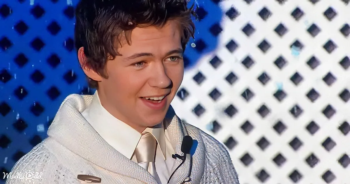 A young man in a white sweater and tie smiles on stage with a microphone, against a blue and white checkered background.