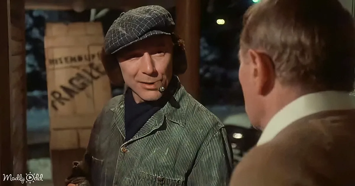A man in a plaid cap and green jacket is speaking to another man in a suit, with a wooden crate labeled "FRAGILE" in the...