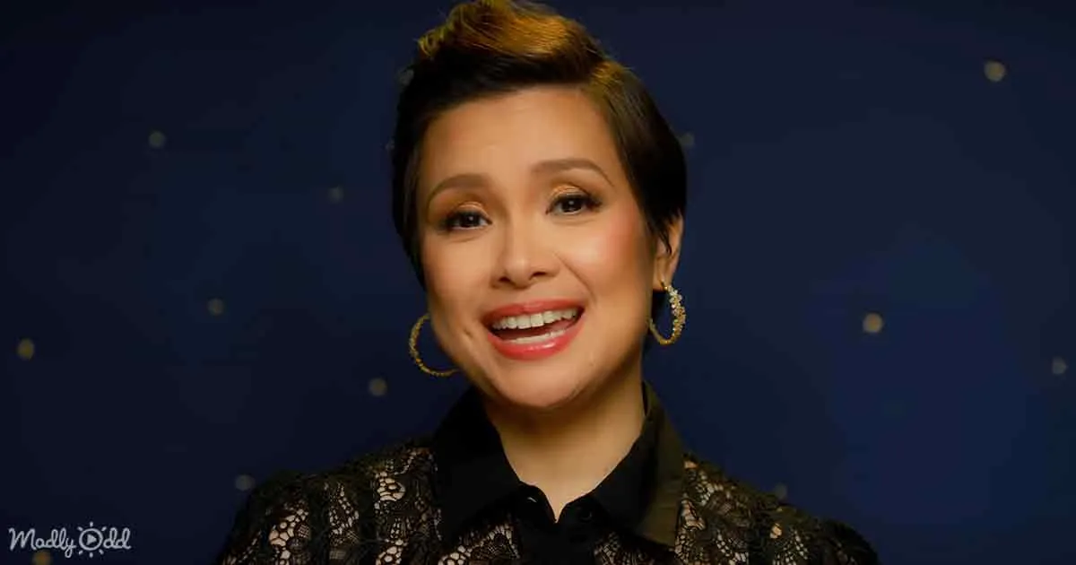 A smiling woman with short hair, wearing a black lace top and gold hoop earrings, against a dark background with small...