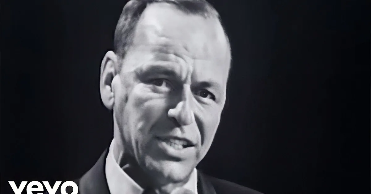 A man in a suit and bow tie speaks into a microphone, captured in black and white.