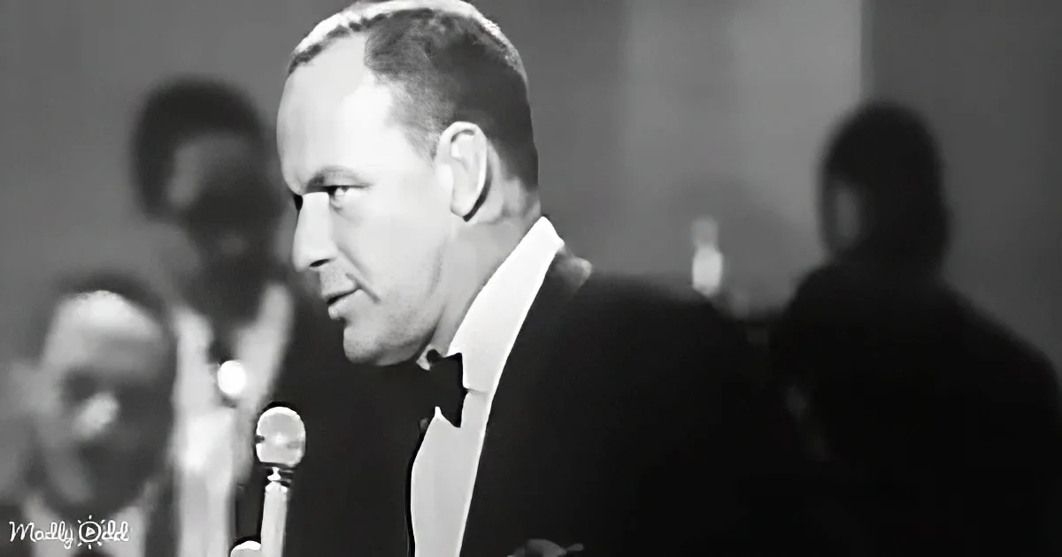 A man in a tuxedo sings into a microphone, with a band in the background.