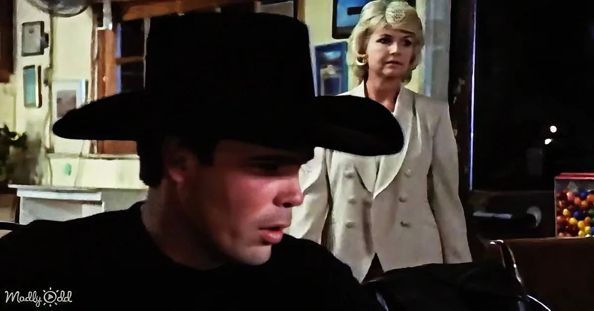 A man in a black shirt and hat sits in a diner, while a woman in a white suit stands behind him.