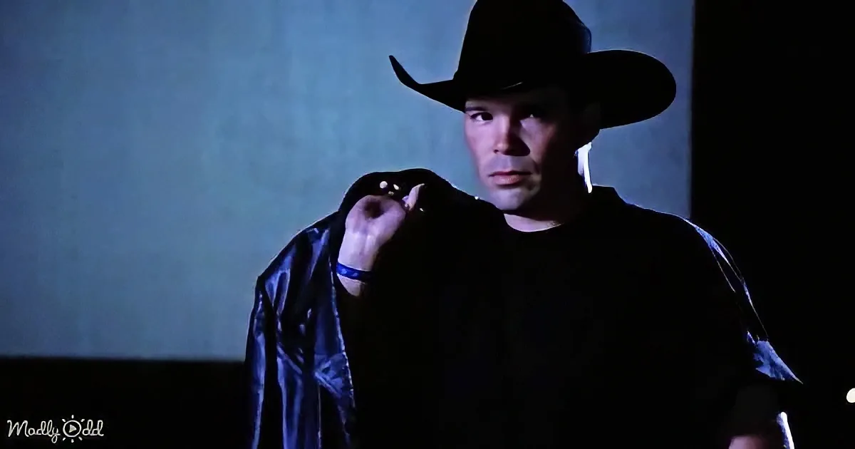 A man in a cowboy hat and dark clothing stands in a dimly lit room, holding his jacket.