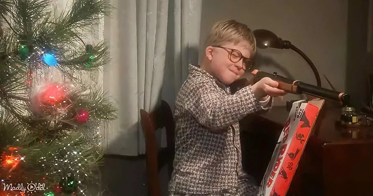 A young boy in a checkered shirt aims a toy rifle near a decorated Christmas tree.
