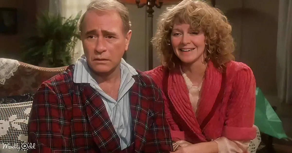A man in plaid pajamas and a woman in a red robe sit together, smiling and looking at the camera.